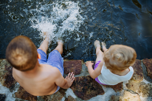 A cute girl and boy sitting together on the footpath by the lake, dangle their feet in the water, summer vacation concept.