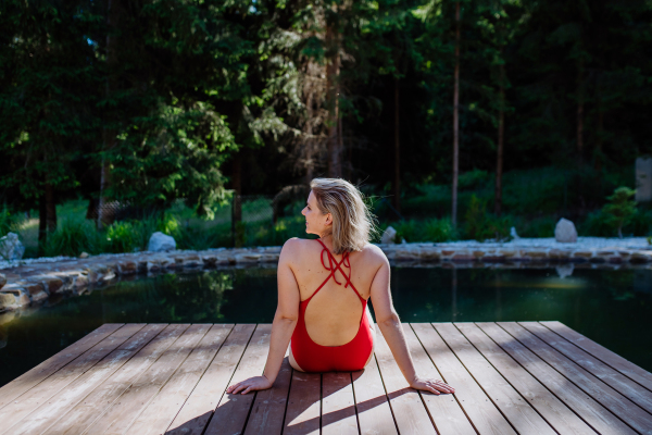 A Rear view of young woman in swimsuit sitting on pier by lake during summer vacation in mountains.