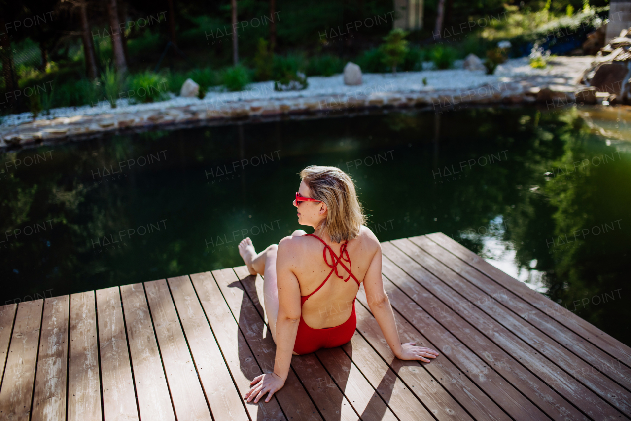 A Rear view of young woman in swimsuit sitting on pier by lake during summer vacation in mountains.