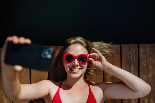 A rlaxed woman taking selfie when lying on a pier by natural lake in summer