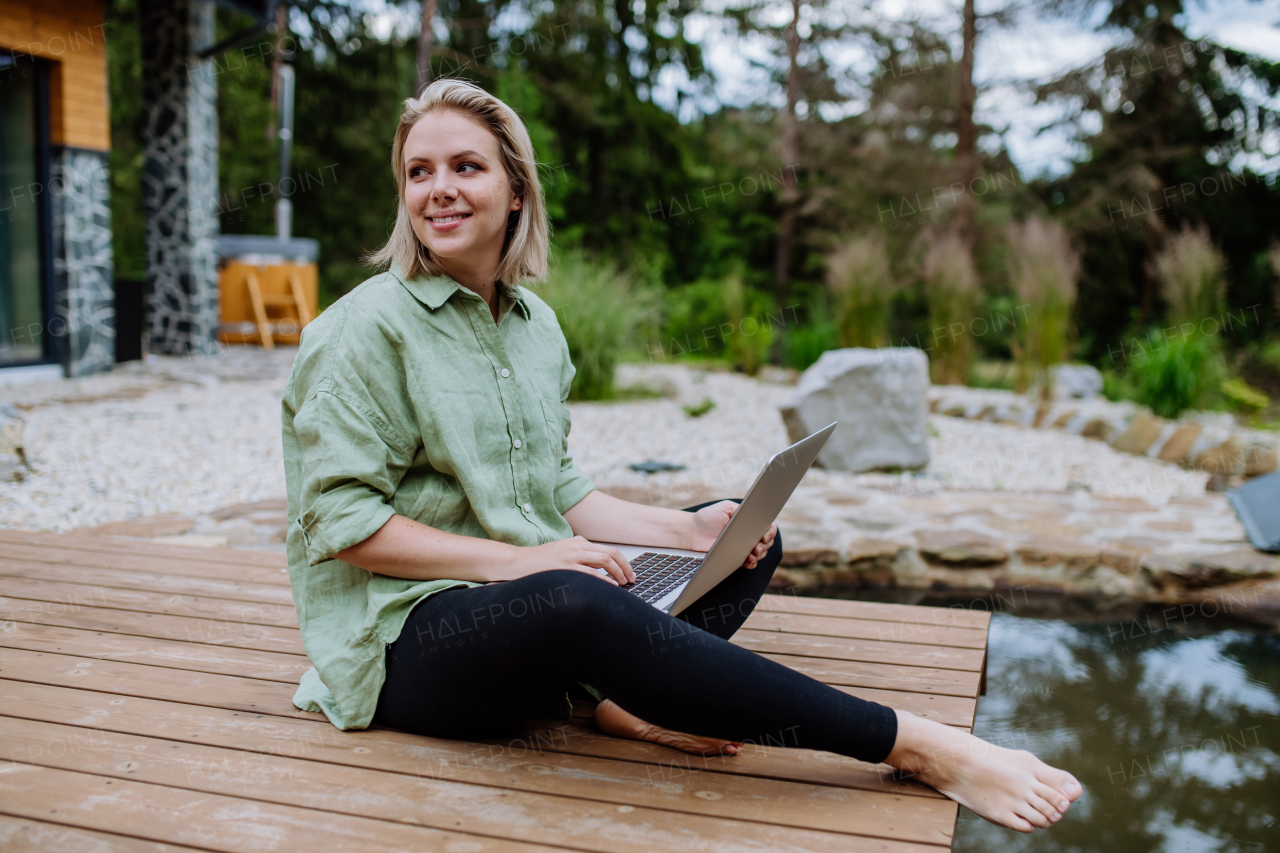 A woman, freelancer working on laptop, sitting on a pier by the backyard lake, a concept of remote office, work during vacation.