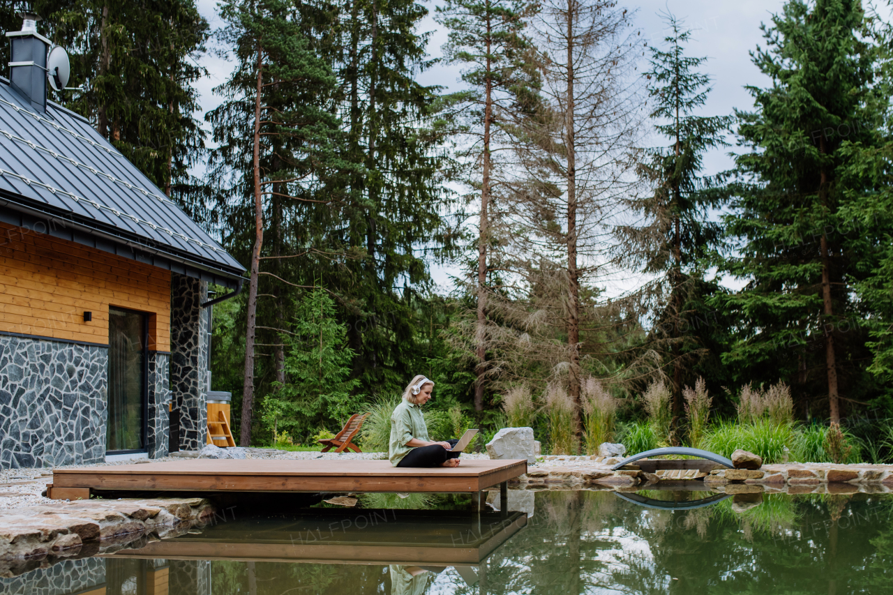 A woman, freelancer working on laptop, sitting on a pier by the backyard lake, a concept of remote office, work during vacation.