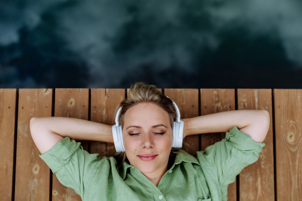 A rlaxed woman wearing headphones listening to music lying on a pier by natureal lake in summer