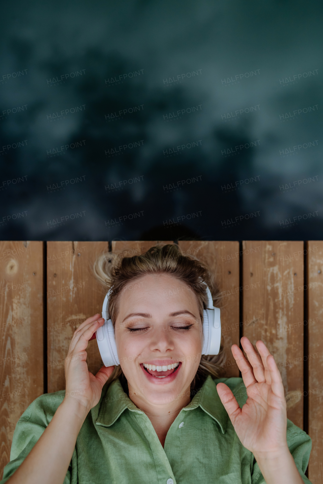 A relaxed woman with headphones listening to music and singing for herself lying on a pier by natureal lake in summer