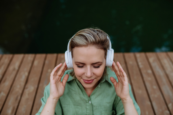 A rlaxed woman wearing headphones listening to music sitting on a pier by natureal lake in summer