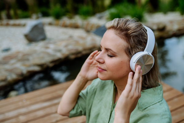 A rlaxed woman wearing headphones listening to music sitting on a pier by natureal lake in summer