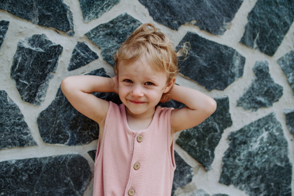 Portrait of little cute girl posing in front of a stone wall, looking at camera.