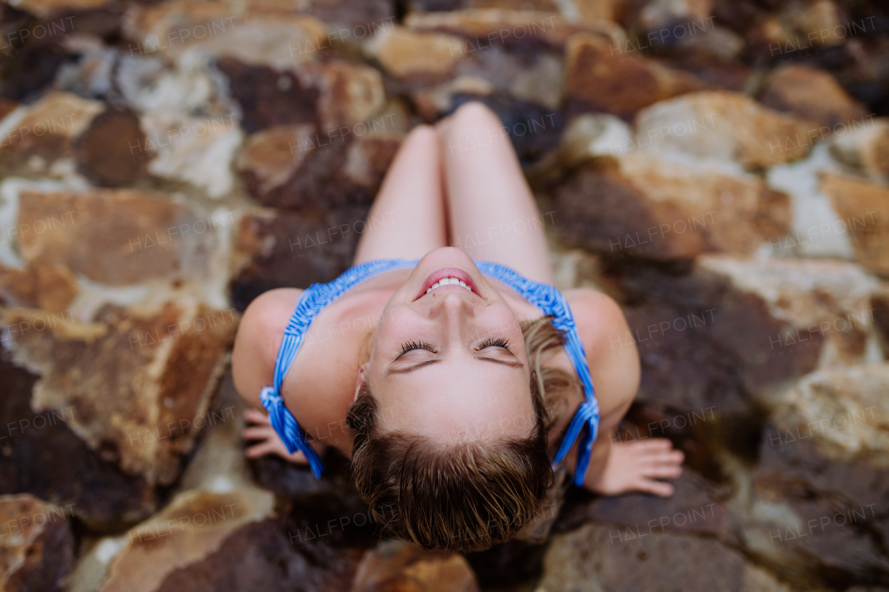 A top view of young woman in swimsuit sitting by backyard natural pond during summer vacation in mountains.