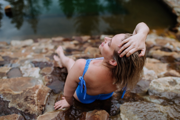 Rear view of happy young woman resting and sunbathing in a swimsuit near lake, during hot sunny day on summer vacation.