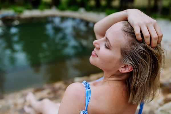 Rear view of happy young woman resting and sunbathing in a swimsuit near lake, during hot sunny day on summer vacation.