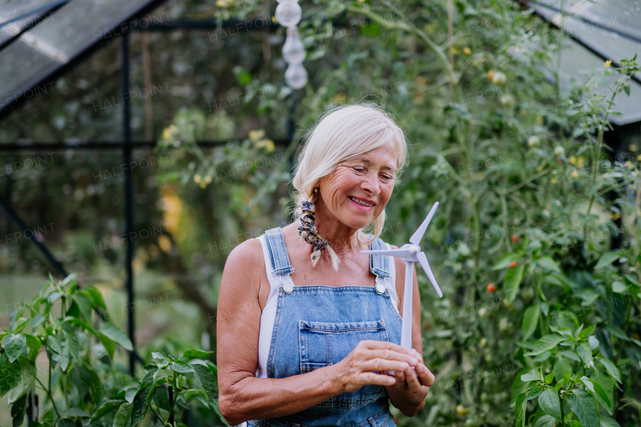 Senior woman holding plastic model of a wind turbine in her greenhouse, concept of renewable resources and eco gardening.