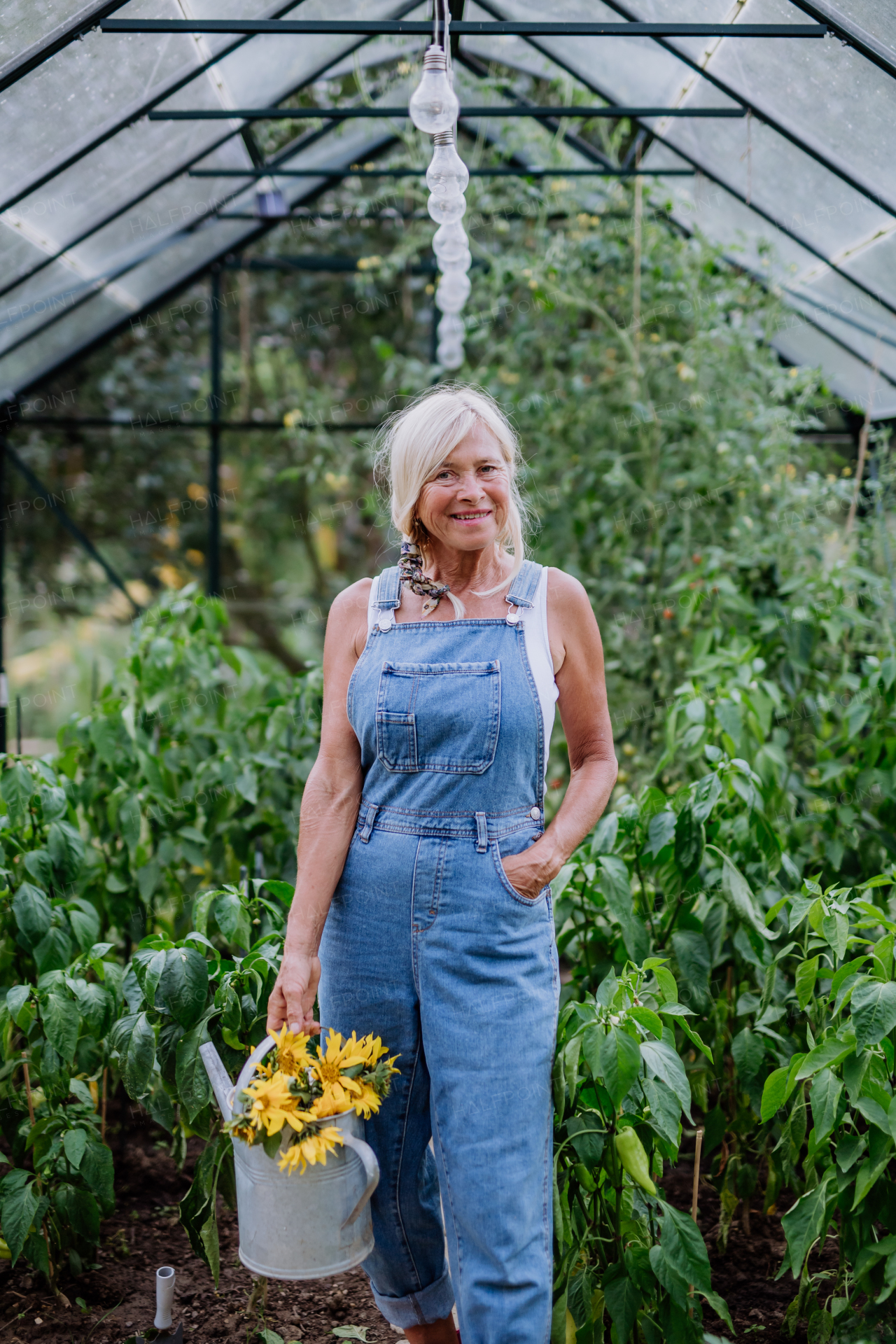 Senior woman posing with a watering pot and flowers in her greenhouse, gardening and sustainable lifestyle concept.