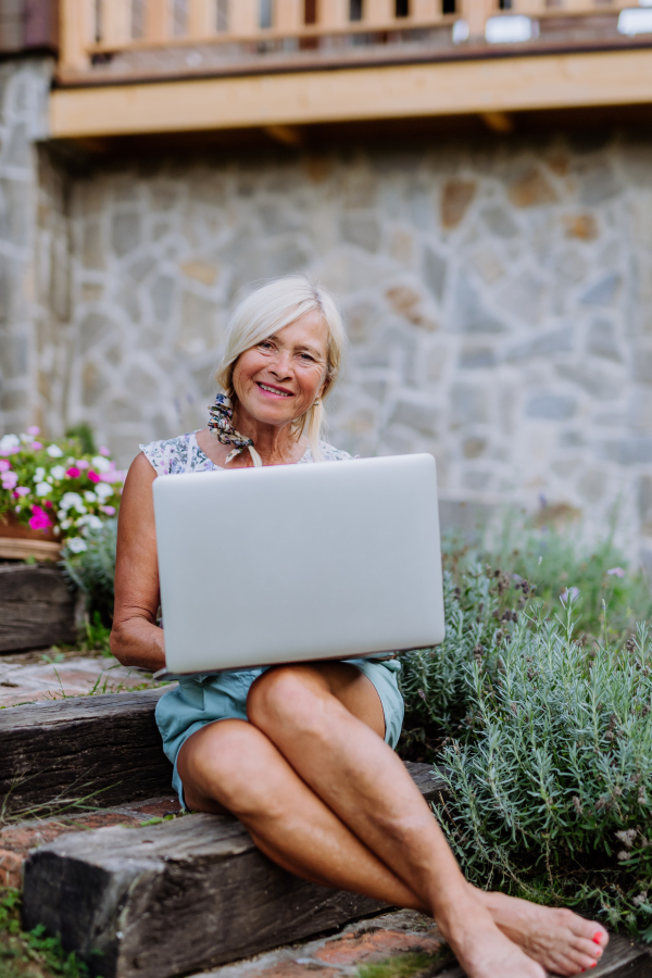 A senior woman using laptop and handling orders of her homegrown organic flowers and vegetables in garden