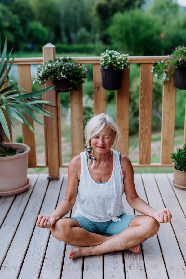 Senior woman sitting outdoors on a terrace in summer, doing yoga exercise.