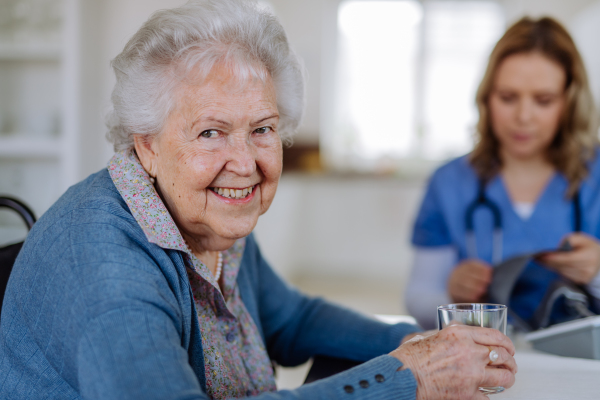 Portrait of senior woman with glass of water, the nurse assistant sitting in background.