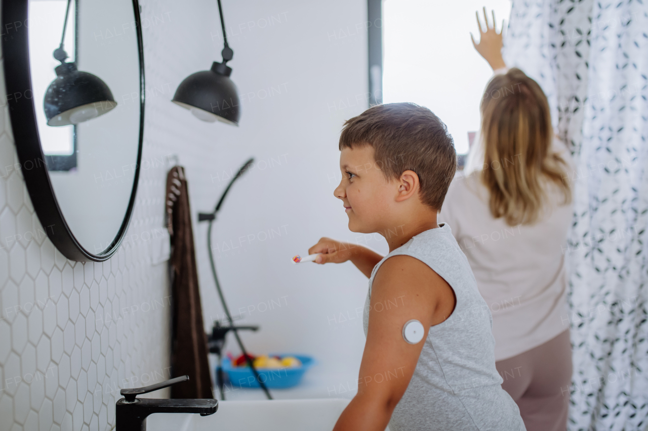 The diabetic boy brushing his teeth in the morning, preparing for the day, while wearing a continuous glucose monitoring sensor on his arm. CGM device making life of school boy easier, helping manage his illness and focus on other activities.