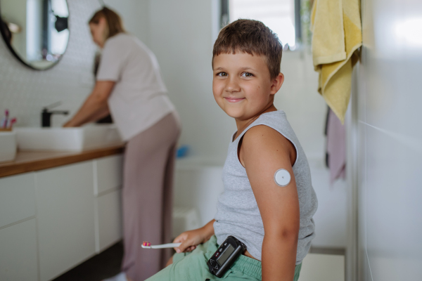 The diabetic boy brushing his teeth in the morning, preparing for the day, while wearing a continuous glucose monitoring sensor on his arm. CGM device making life of school boy easier, helping manage his illness and focus on other activities.