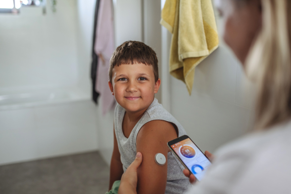 Boy with diabetes checking blood glucose level at home using continuous glucose monitor. The boy's mother connects his CGM to a smartphone to monitor his blood sugar levels in real time.