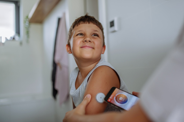 Boy with diabetes checking blood glucose level at home using continuous glucose monitor. The boy's mother connects his CGM to a smartphone to monitor his blood sugar levels in real time.