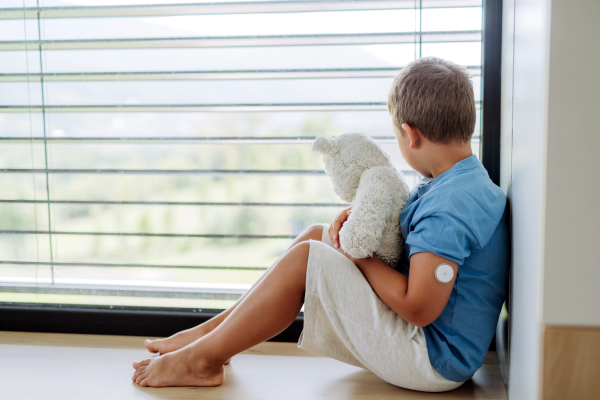 Diabetic boy with a continuous glucose monitor sitting by the window, holding his stuffed teddy bear and looking outside. Children with diabetes feeling different or isolated from peers.