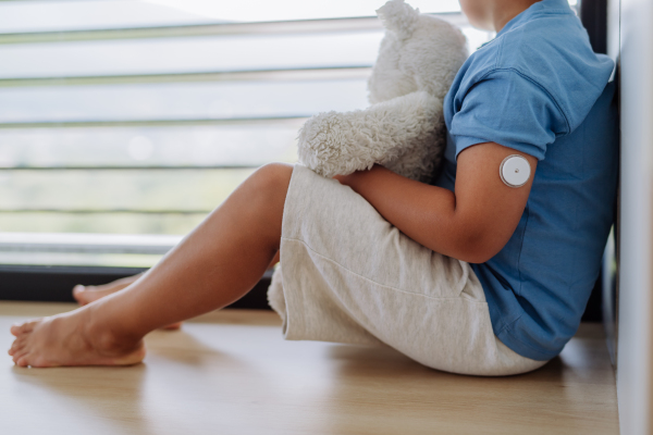 Diabetic boy with a continuous glucose monitor sitting by the window, holding his stuffed teddy bear and looking outside. Children with diabetes feeling different or isolated from peers.