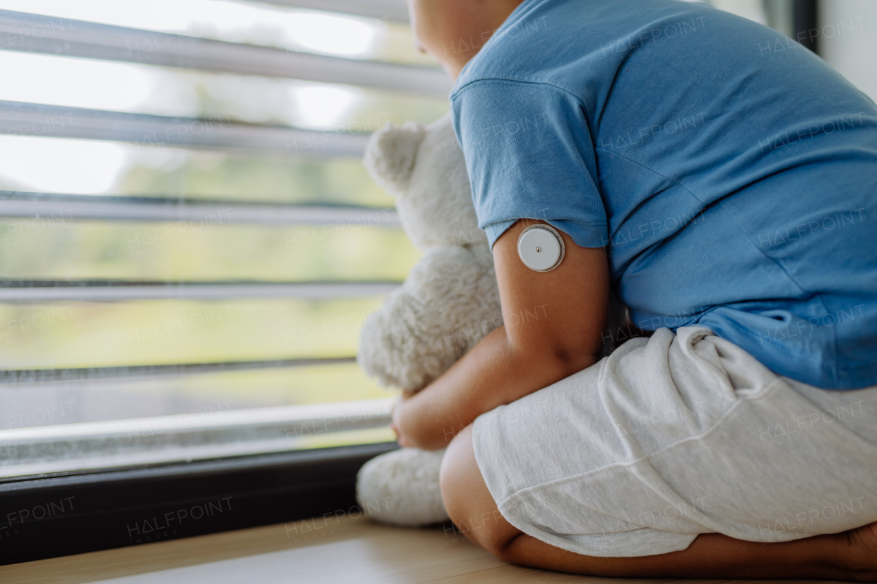 Diabetic boy with a continuous glucose monitor sitting by the window, holding his stuffed teddy bear and looking outside. Children with diabetes feeling different or isolated from peers.