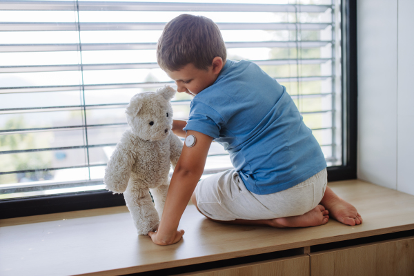 Diabetic boy with a continuous glucose monitor sitting by the window, showing his stuffed teddy bear sensor on his arm. Children with diabetes feeling different or isolated from peers.