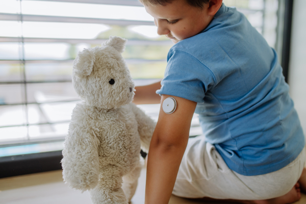 Diabetic boy with a continuous glucose monitor sitting by the window, showing his stuffed teddy bear sensor on his arm. Children with diabetes feeling different or isolated from peers.