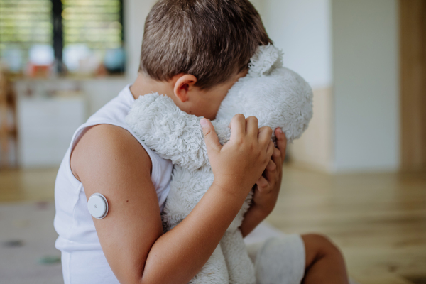 Diabetic boy with a continuous glucose monitor sitting at home, hugging his stuffed teddy bear. Children with diabetes feeling different or isolated from peers.