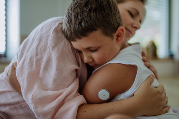 Diabetic boy with a continuous glucose monitor embracing his mother, feeling sad. Children with diabetes feeling different or isolated from peers.