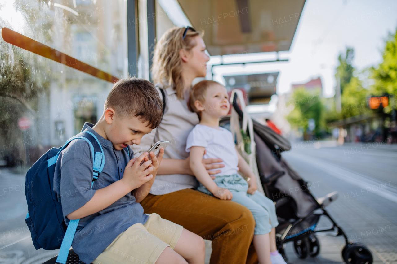 A young mother with little kids waiting on bus stop in city, scrolling on mobile phone.