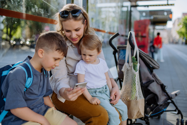 A young mother with little kids waiting on bus stop in city, scrolling on mobile phone.