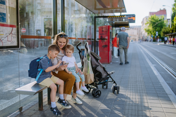 A young mother with little kids waiting on bus stop in city, scrolling on mobile phone.