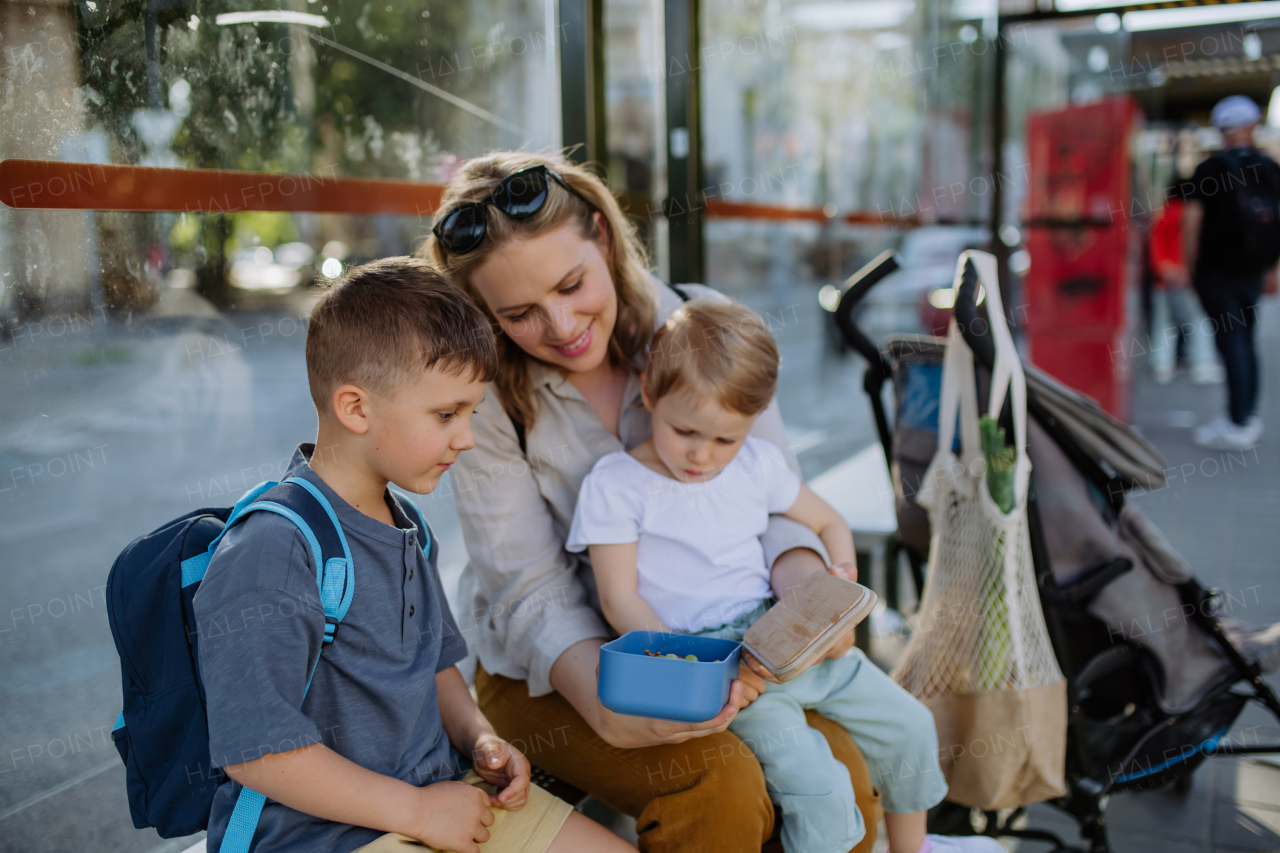 A young mother with little kids waiting on bus stop in city and eating fruit snack.