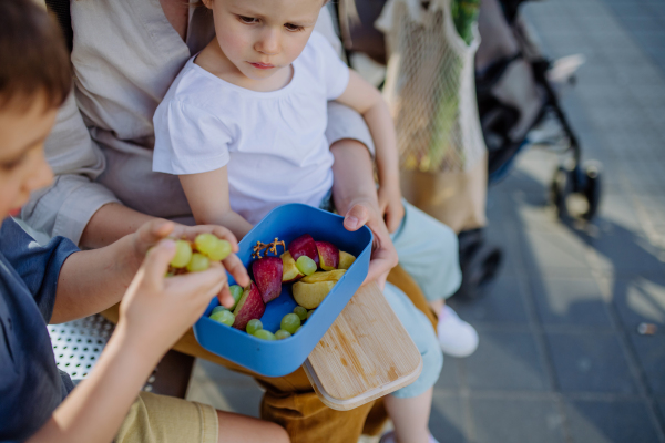 A young mother with little kids waiting on bus stop in city and eating fruit snack.