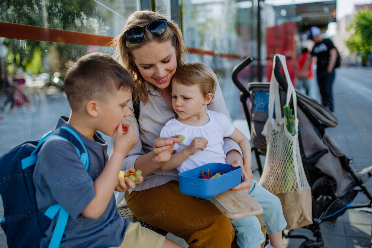 A young mother with little kids waiting on bus stop in city and eating fruit snack.