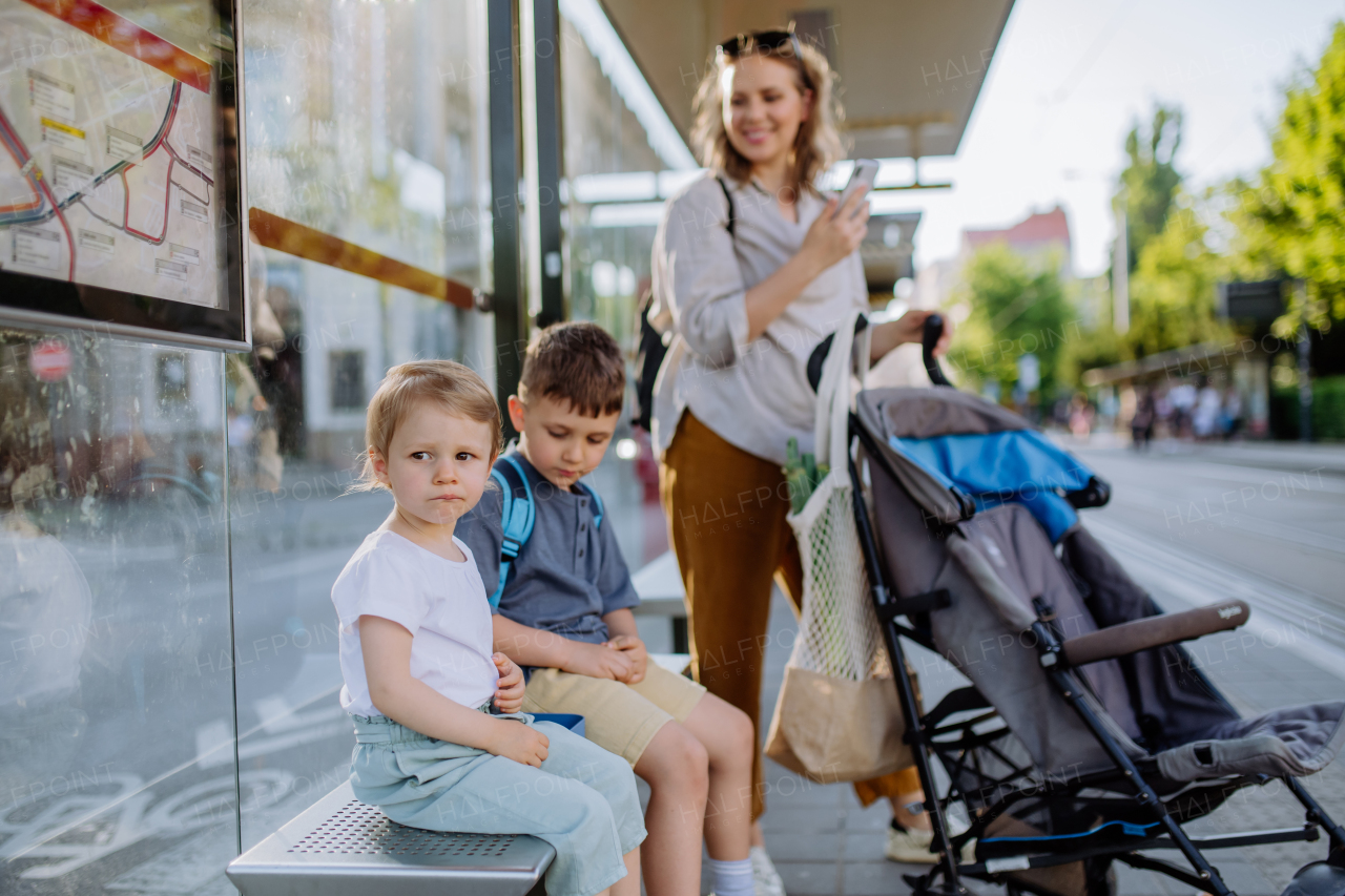 A young mother with little kids waiting on bus stop in city.