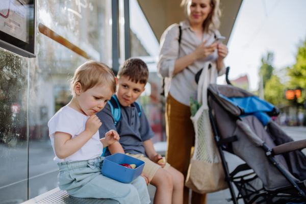 A young mother with little kids waiting on bus stop in city and eating fruit snack.