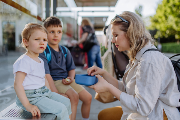 A young mother with little kids waiting on bus stop in city and eating fruit snack.