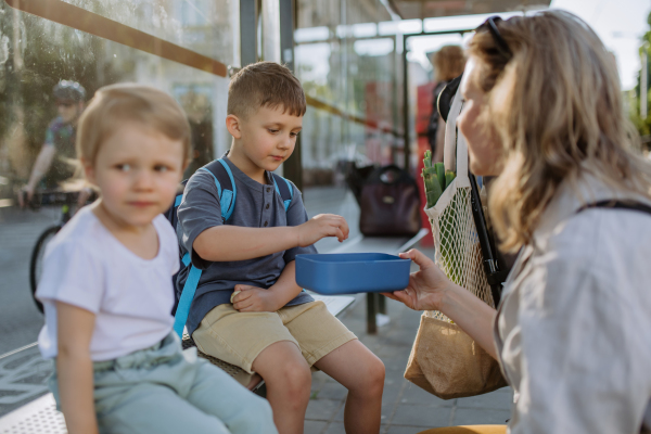 A young mother with little kids waiting on bus stop in city and eating fruit snack.