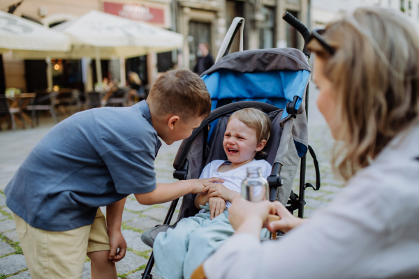 A mother and little brother calming a crying girl sitting in a stroller while walking down the street