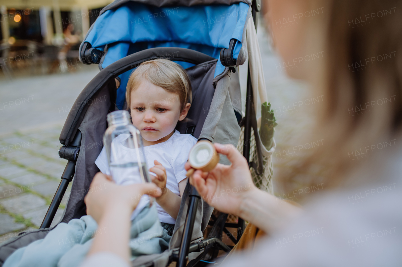 An unrecognizable mother holding zero waste water bottle and opening it in front of her little child in stroller, sustainable lifestyle.