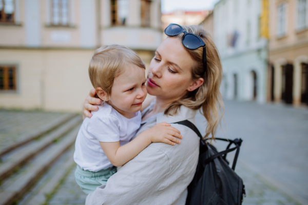 A mother consling her little daughter crying, holding her in arms in street in summer.