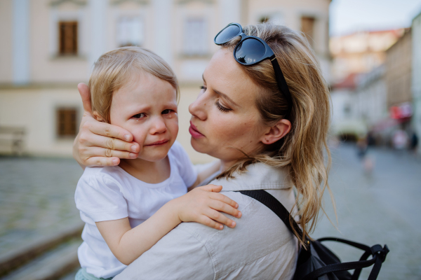 A mother consling her little daughter crying, holding her in arms in street in summer.