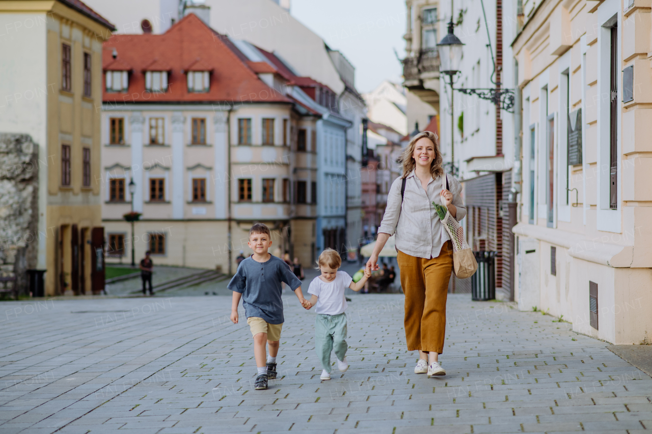 A young mother with zero waste shopping bag holding hands with her children and walking in old city street center.