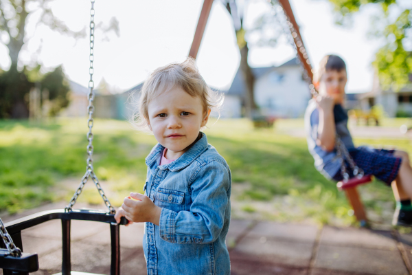 Little sibling playing together in playground, swaying on a swing, enjoying sunny summer day.