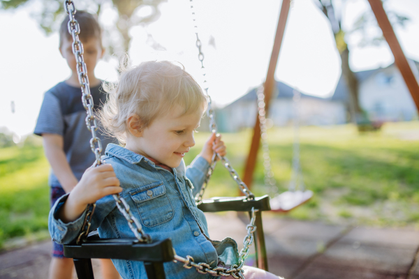 Little sibling playing together in playground, swaying on a swing, enjoying sunny summer day.