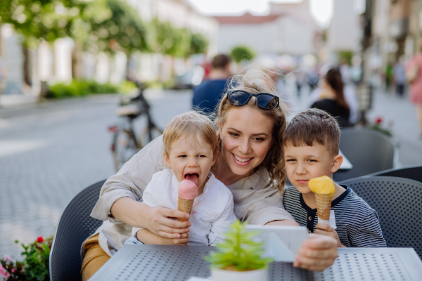 A young mother taking selfie with her kids eating ice-cream in cafe outdoors in street in summer.