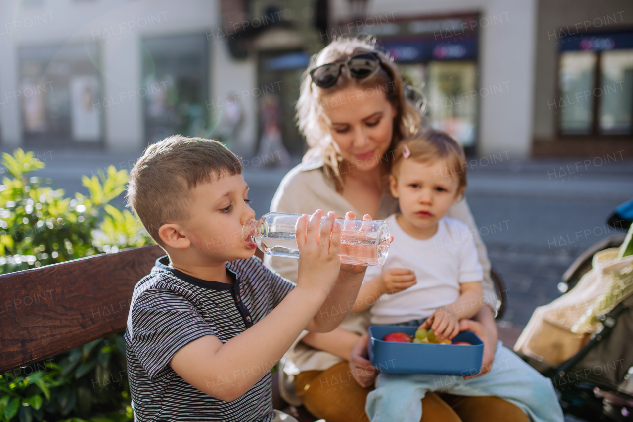 A young mother with little kids sitting on bench in city in summer, eating fruit snack and drinking water.