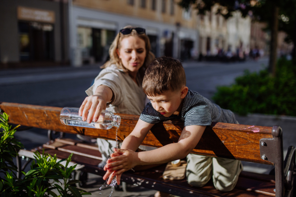 A young mother holding zero waste water bottle and washing hands of her little sonwhen sitting on bench in city, sustainable lifestyle.